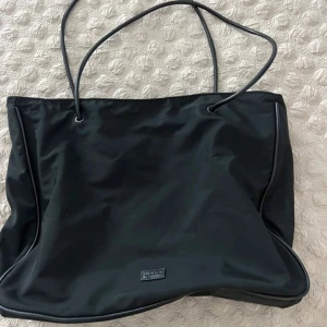 Svart shoppingväska i syntet - Snygg och praktisk svart. Perfekt för vardagsbruk med gott om plats för alla dina saker. Väskan har långa handtag som gör den enkel att bära över axeln. En stilren och tidlös design som passar till alla tillfällen.