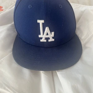 Blå keps från New Era - Säljer en snygg blå keps från New Era med LA-logga framtill. Kepsen är i bra skick och har en klassisk 59FIFTY passform. Perfekt för sommarens alla äventyr eller för att visa ditt stöd för ditt favoritlag!