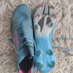 Blå fotbollsskor från Adidas - Säljer ett par snygga blå fotbollsskor från Adidas i mycket bra skick. De har rosa detaljer och snörning för perfekt passform. Skorna är designade för att ge bra grepp på planen med sina dobbar. Perfekta för fotbollsträning eller match!
