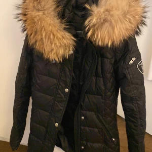 Svart quiltad jacka med pälsdetalj - Säljer en snygg svart quiltad jacka från Rockandblue med en stor, fluffig pälsdetalj på kragen. Jackan har långa ärmar och en dragkedja framtill med knappar. Perfekt för kalla vinterdagar och höstpromenader. Den har en cool patch på ärmen som ger en extra touch av stil.
