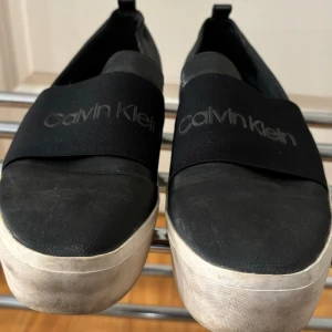 Svarta loafers från Calvin Klein - Säljer ett par svarta loafers från Calvin Klein i bra skick. De har en elastisk rem med logotyp över foten och en vit sula som ger en snygg kontrast. Perfekta för både vardag och fest!