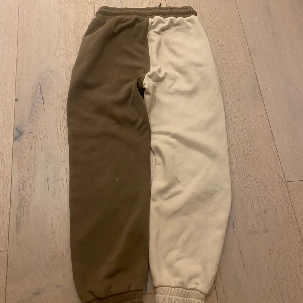 Säljer ett par coola mjukisbyxor från Kappahl i storlek 116. Byxorna har en unik design med en ben i beige och det andra i brunt, samt svarta detaljer. De har elastisk midja med snörning och dragkedjor på fickorna. Perfekta för en avslappnad stil!. Jeans & Byxor.