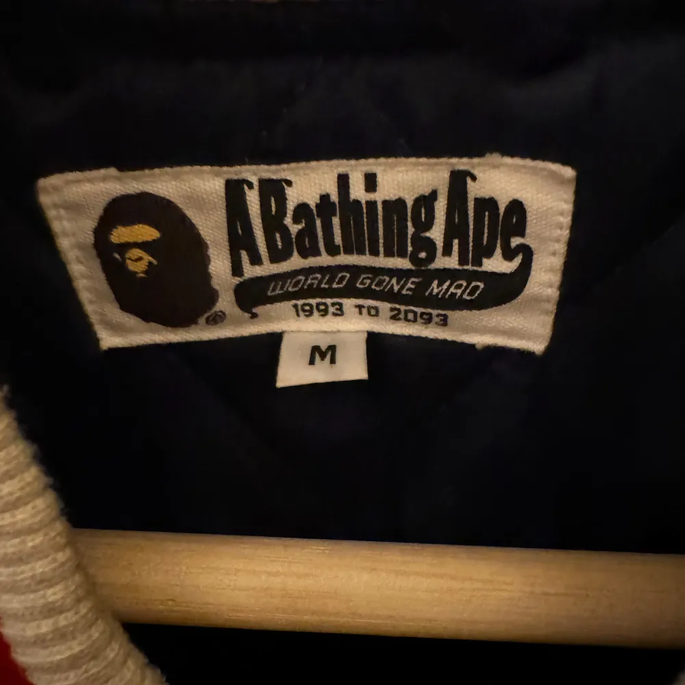 Säljer en snygg mörkblå bomberjacka från BAPE med vita och röda detaljer. Jackan har ett coolt tryck med texten 'A Bathing Ape' på ryggen och 'BAPE' på framsidan. Den är quiltad inuti för extra komfort och värme. Perfekt för höst och vår!. Jackor.