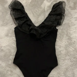 Svart body från Amisu - Snygg svart body från Amisu med volangdetaljer på axlarna. Perfekt för en utekväll eller fest. Den har en elegant och stilren design som passar till många olika outfits. Väldigt trendig!