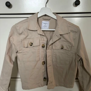 Beige jacka från Lindex - Säljer en snygg beige jacka från Lindex i storlek 140. Jackan har långa ärmar och knappar framtill samt två bröstfickor med knappar. Den högra knappen är halftsönder. Perfekt för vår och höst! 🧥