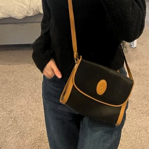 YSL Svart och brun axelväska - Snygg svart axelväska med bruna detaljer och justerbar rem. Väskan har en stilren design med ett emblem på framsidan och praktisk knäppning. Perfekt för vardagsbruk eller en utekväll. Den har ett huvudfack med dragkedja för säker förvaring. Knappt använd och är i nyskick.