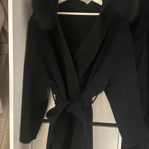 Svart kappa med pälsdetaljer - Säljer en elegant svart kappa med pälsdetaljer på kragen (fuskpäls). Den har ett knytbälte i midjan för en snygg passform. Perfekt för kyliga höst- och vinterdagar. Kappan är långärmad och ger en stilren look.
