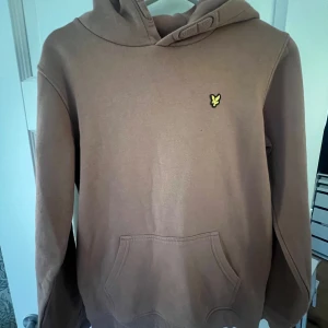 Brun hoodie med logga - Säljer en brun Lyle Scott hoodie i bra skick med en gul logga på bröstet. Den har en klassisk känguruficka och en bekväm huva. Perfekt för höst och vinter, och passar till vardags eller chillkvällar. Skön och stilren design!