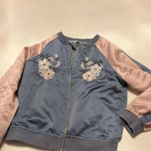 Blå och rosa bomberjacka från Lindex - Säljer en superfin blå och rosa bomberjacka från Lindex i storlek 146/152. Jackan har snygga broderade blommor och en fågel på både fram- och baksidan. Den har en dragkedja i guld och ribbade muddar. Perfekt för vår och höst!