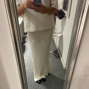 Vit långkjol - Säljer en superfin vit ribbad långkjol i stickat material. Kjolen har en elegant slits på sidan. Den är i mycket bra skick och passar perfekt till höst- och vintergarderoben. Kjolen är bekväm och stilren, vilket gör den lätt att matcha med.