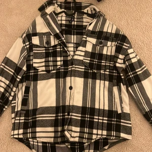 Svartvit rutig jacka - Säljer en snygg svartvit rutig jacka med knappar framtill. Den har två bröstfickor med knappar och en skön huva. Perfekt för höst och vår när du vill hålla stilen. Jackan är i bra skick och passar både till vardags och lite mer avslappnade tillfällen.