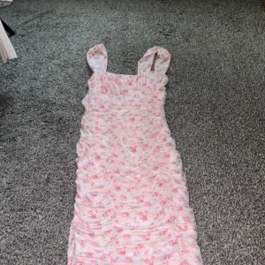 Rosa blommig klänning - Söt rosa klänning med blommigt mönster, perfekt för sommarens fester. Den har en tight passform och ärmlös design med breda axelband. Materialet känns lätt och luftigt, vilket gör den idealisk för varma dagar. Klänningen är i mycket bra skick och redo att bäras till nästa fest eller midsommarfirande.