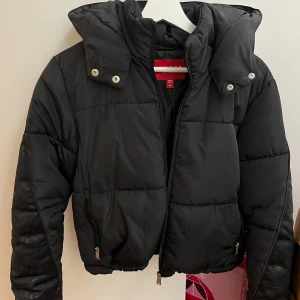Svart pufferjacka från Guess - Säljer en snygg svart pufferjacka från Guess i storlek M. Jackan har en cool quiltad design med dragkedja och knappar. Perfekt för höst och vinter, håller dig varm och stilren. Den har en normal passform och är i mycket bra skick. Passar perfekt för både vardag och fest!