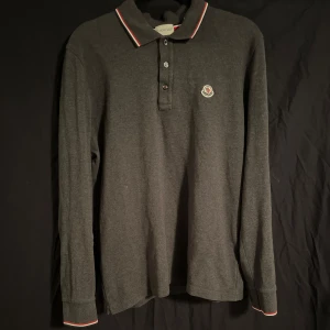 Grå pikétröja från Moncler - Säljer en snygg grå pikétröja från Moncler i mycket bra skick. Tröjan har långa ärmar och en klassisk krage med knappar. Den har Monclers logga broderad på bröstet och detaljer i rött och vitt på kragen och ärmsluten. Perfekt för både vardag och fest! Storlek L men sitter som mer än M.