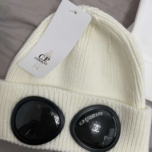  C.P. Company mössa  - Säljer en supercool vit stickad mössa från C.P. Company. Den har de ikoniska glasögondetaljerna framtill som ger en unik look. Perfekt för att hålla sig varm och stilren under kalla dagar. Passar perfekt till vinterns alla outfits! Helt oanvända 