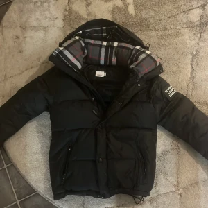 Svart dunjacka från Burberry - Säljer en snygg svart dunjacka använd några gånger från Burberry med klassiskt rutigt foder i huvan. Kan ej bli vest det är annan model Jackan har en dragkedja och knappar framtill, perfekt för kalla vinterdagar. Den är i mycket bra skick och har en stilren design med Burberry-logga på ärmen. Passar perfekt för både vardag och fest!