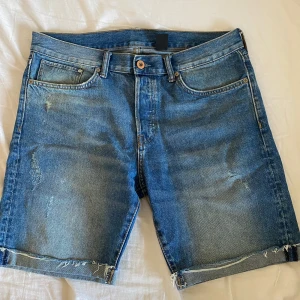 Blå jeansshorts - Säljer ett par snygga blå jeansshorts i straight fit. De har en klassisk femficksdesign och slitna detaljer för en cool look. Perfekta för sommaren och avslappnade dagar. Shortsen är i bra skick och gjorda av denim.
