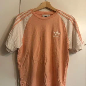 Persikofärgad t-shirt från Adidas - Säljer en snygg persikofärgad t-shirt från Adidas med vita och rosa ränder på ärmarna. Den har en klassisk Adidas-logga på bröstet och är i ett mjukt bomullsmaterial. Perfekt för en avslappnad stil eller träning. Passar bra till både jeans och träningsbyxor!