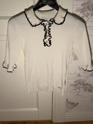 Vit ribbad topp med volangkrage Holly & Whyte - Supersöt vit ribbad topp från Holly & Whyte med volangkrage och svarta kontrastkanter. Knappar framtill med pärldetaljer och volang vid ärmslut. Perfekt till jeans eller kjol för en preppy look.