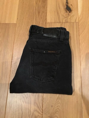 Svarta jeans från Nudie Jeans - Säljer ett par svarta jeans från Nudie Jeans med klassisk femficksdesign och diskret logga på bakfickan. Jeansen har raka ben och normal passform, tillverkade i slitstark bomull. Perfekta för dig som gillar stilrena och tidlösa plagg.