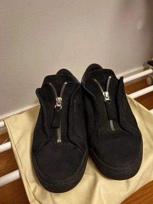 Svarta sneakers från Axel Arigato - Säljer ett par svarta sneakers från Axel Arigato i mocka med dragkedja framtill och diskret guldfärgad logga på sidan. Skorna har rund tå, platt sula och detaljer i svart skinn på hälen. Snygg minimalistisk design som passar till det mesta. Innersulan är lite trasig, se bild 5, men går att byta ut om man skulle vilja. Original dustbag och låda medföljer!