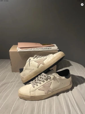 Golden Goose St43 - Golden Goose Superstar sneakers i vitt skinn med grå mockastjärna på sidan, grå snörning och svart häl. Skorna är riktigt bra skick förutom ett litet hål i hälen. Därav priset. Se bild. Snygga med bra kontrast mellan färgerna och den ikoniska stjärndetaljen som gör dem riktigt trevliga nu till hösten och sommaren. Storleken är 43. Hör av er vid eventuella frågor.