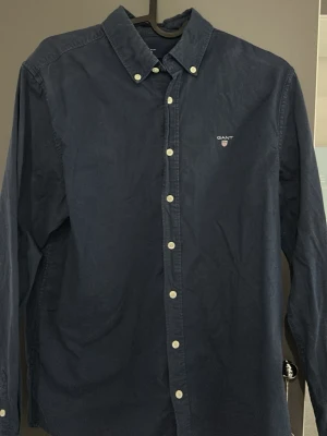 Mörkblå skjorta från Gant - Klassisk mörkblå skjorta från Gant med button-down krage och vita knappar. Skjortan har lång ärm och diskret Gant-logga broderad på bröstet. Tillverkad i mjuk bomull för en skön känsla och snygg passform. Storleken är 15 år/170 cm men skulle säga att den sitter som en XS men fråga gärna om du är osäker! Pris går att diskutera!