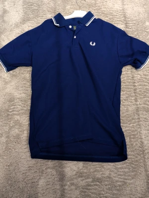 Marinblå pikétröja från Fred Perry - Säljer en klassisk marinblå pikétröja från Fred Perry med vit logga broderad på bröstet. Tröjan har vit rand på krage och ärmslut, samt knappar vid halsen. Perfekt till en avslappnad men ändå stilren look.