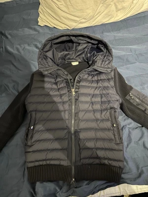 Marinblå Moncler pufferjacka med huva - Säljer en marinblå pufferjacka från Moncler med stickade ärmar och huva. Jackan har dragkedja framtill, snörning i huvan och ribbade muddar. Klassisk Moncler-logga på ärmen och praktiska fickor med knappstängning. Perfekt för kyliga dagar.