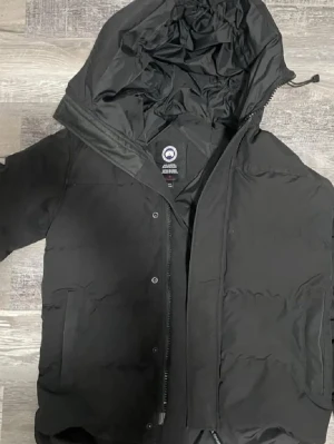 Svart dunjacka från Canada Goose - Säljer en svart dunjacka från Canada Goose med huva och dragkedja. Jackan har en klassisk look med diskret logga på ärmen och praktiska fickor framtill. Perfekt för kalla dagar och riktigt snygg till streetwear-stilen.