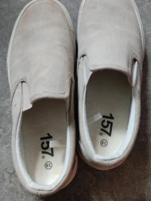 Beige slip-on sneakers från 157 - Säljer ett par beige slip-on sneakers från 157 i storlek 38. Skorna har en klassisk, enkel design med vit sula och ovandel i canvas. De är lätta att ta på och av tack vare elastiska paneler på sidorna. Perfekta för en chill och avslappnad stil.