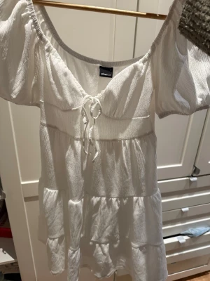 Vit kortklänning från Gina Tricot - Supersöt vit kortklänning från Gina Tricot med puffärmar och knyt i fram. Klänningen har en luftig, rynkad struktur och volangdetaljer i flera lager. Perfekt för varma dagar och ger en romantisk vibe.