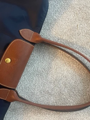 Marinblå axelväska från Longchamp - Säljer en klassisk marinblå axelväska från Longchamp med bruna skinnhandtag och detaljer. Väskan har guldfärgade metalldetaljer och dragkedja. Perfekt storlek för att få plats med allt du behöver och enkel att bära över axeln.