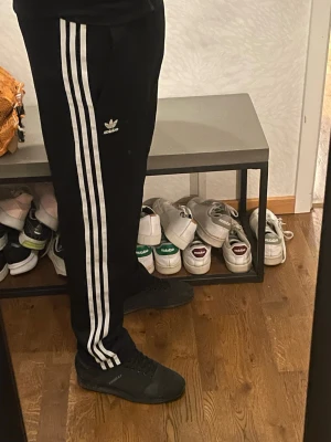 Svarta Adidas byxor med vita ränder - Svarta Adidas byxor med klassiska tre vita ränder längs sidorna och Adidas-logga på låret. Byxorna har en normal passform och resår i midjan. Perfekta för en sportig look eller chill vardagsstil.