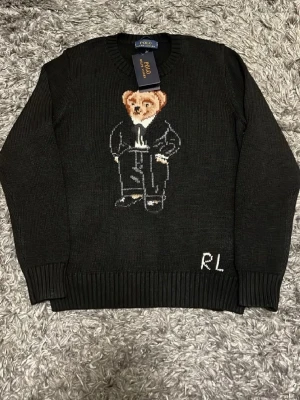 Svart stickad tröja Polo Bear RL - Svart stickad tröja från Polo Ralph Lauren med ikoniska Polo Bear-motivet framtill och RL-broderi vid nederkanten. Tröjan har rund hals, ribbade muddar och är långärmad. Perfekt för dig som vill ha en klassisk men lekfull look.(Säljer för att den var för liten.)