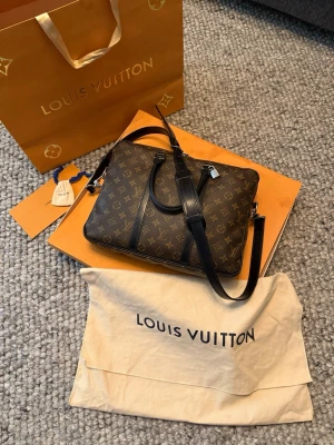 Louis Vuitton portfölj - Säljer en klassisk Louis Vuitton datorväska i brunt med ikoniskt monogrammönster och svarta skinnhandtag. Väskan har avtagbar axelrem, silverfärgade metalldetaljer och dragkedja. Perfekt för dig som vill ha en lyxig och stilren accessoar till vardagen.