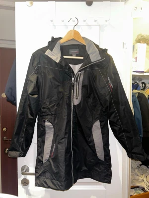 Svart vindjacka från Autograph XS - Säljer en svart vindjacka från Autograph i storlek XS. Jackan har gråa detaljer, huva med dragsko och flera fickor med dragkedja. Materialet är lätt och känns sportigt, perfekt för blåsiga dagar. Snygg och funktionell design med reflekterande detaljer.