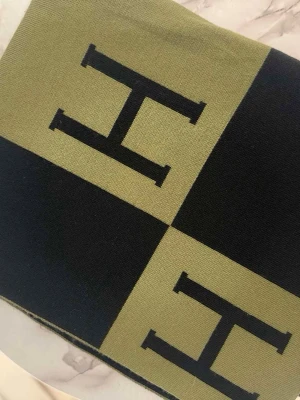 Beige och svart stickad väst med H-mönster - Säljer en snygg stickad väst i beige och svart med stora H-tryck i rutigt mönster. Västen har en modern och grafisk look som sticker ut. Perfekt för dig som vill ha något unikt och trendigt i garderoben.