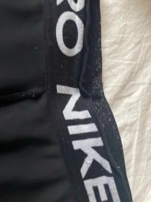 Svarta Nike Pro tights - Säljer ett par svarta Nike Pro tights med vit logga på benet och bred resår i midjan med Nike Pro-tryck. Sitter tight och är perfekta för träning eller gymmet. Materialet är stretchigt och följsamt för maximal rörelsefrihet. De är lite slitna!