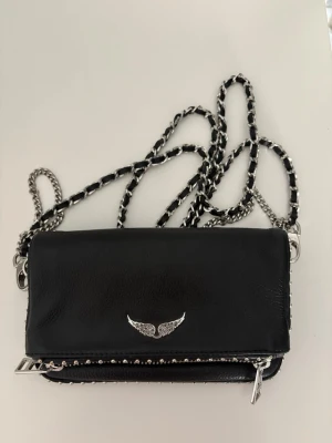 Svart axelväska Zadig & Voltaire  - Snygg svart axelväska från Zadig & Voltaire i skinn med silverfärgade kedjedetaljer och ikonisk vinge framtill. Väskan har dragkedja, nitar runt kanterna och justerbar kedjerem. Perfekt storlek för det viktigaste och ger en cool vibe till din outfit. Mått: 18x11cm Nypris:2700-3300