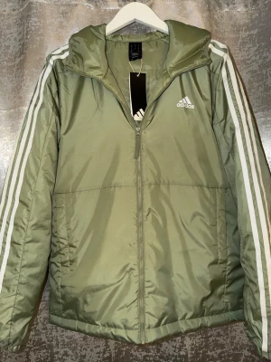 Adidas jacka - Snygg grön pufferjacka från Adidas med huva och klassiska vita ränder längs ärmarna. Jackan har dragkedja framtill, två sidofickor och Adidas-logga på bröstet. Tillverkad i polyester och perfekt för kyligare dagar.