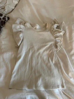 Ribbad vit topp med volangärmar - Säljer en ribbad vit topp med stora volangärmar. Toppen har rund halsringning och är tillverkad i ett mjukt, stretchigt material som känns skönt mot huden. Perfekt för dig som vill ha något extra till outfiten och gillar voluminösa detaljer.