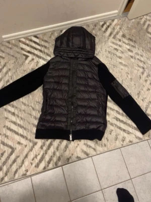 Svart Moncler dunjacka med huva - Säljer en svart Moncler dunjacka med huva och dragkedja framtill. Jackan har quiltad kropp i dun och släta, stickade ärmar med ficka på ena ärmen. Perfekt för dig som vill ha en stilren och varm jacka med exklusiv känsla.
