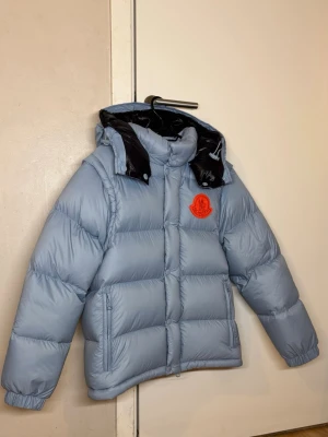 Moncler pufferjacka - Säljer en ljusblå pufferjacka från Moncler med avtagbara ärmar så den kan bäras som väst. Jackan har svart innerfoder, stor huva och röd Moncler-logga på bröstet. Stängs med dragkedja och tryckknappar, samt har två fickor med dragkedja framtill.