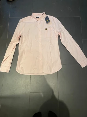 Ljusrosa skjorta från Lyle & Scott - Säljer en ljusrosa skjorta från Lyle & Scott med klassisk krage, bröstficka och den ikoniska gula loggan på bröstet. Skjortan har knappar hela vägen och långa ärmar. Perfekt för dig som vill ha en stilren och enkel look.