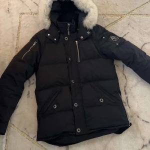Svart dunjacka med päls från Moose Knuckles - Säljer en svart dunjacka från Moose Knuckles med stor huva och vit pälsdetalj. Jackan har flera dragkedjor, tryckknappar och logga på ärmen. Perfekt för kalla dagar och har en riktigt schysst oversized look den är äkta och bra kvaliteé