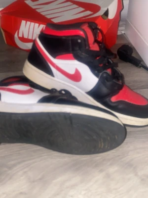 Nike Air Jordan 1 svart/röd/vit - Säljer ett par Nike Air Jordan 1 med klassisk svart, röd och vit färgkombination. Skorna har högt skaft, snörning och den ikoniska swoosh-loggan i rött på sidan. Tillverkade i skinn med vit mellansula och svart yttersula. Perfekta för dig som gillar streetwear och sneakers.