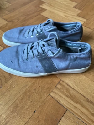 Grå sneakers från Polo Ralph Lauren - Säljer ett par stilrena grå sneakers från Polo Ralph Lauren. Skorna har snörning, vit platt sula och detaljer i mocka på hälen. Tillverkade i bomullstyg med diskret logga på sidan. Perfekta till jeans eller shorts för en avslappnad look.
