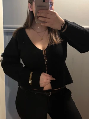 Svart kofta med kedjeknappar - Säljer en svart figursydd kofta med unika kedjeknappar framtill. Tröjan har djup v-ringning och långa ärmar, perfekt för dig som vill ha en stilren men edgy look. Passar till både jeans och kjol för en trendig outfit.