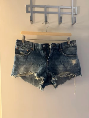 Blå slitna jeansshorts - Säljer ett par skit snygga låg midjade  blå jeansshorts med råa, fransiga kanter och slitningar från Gina tricot 